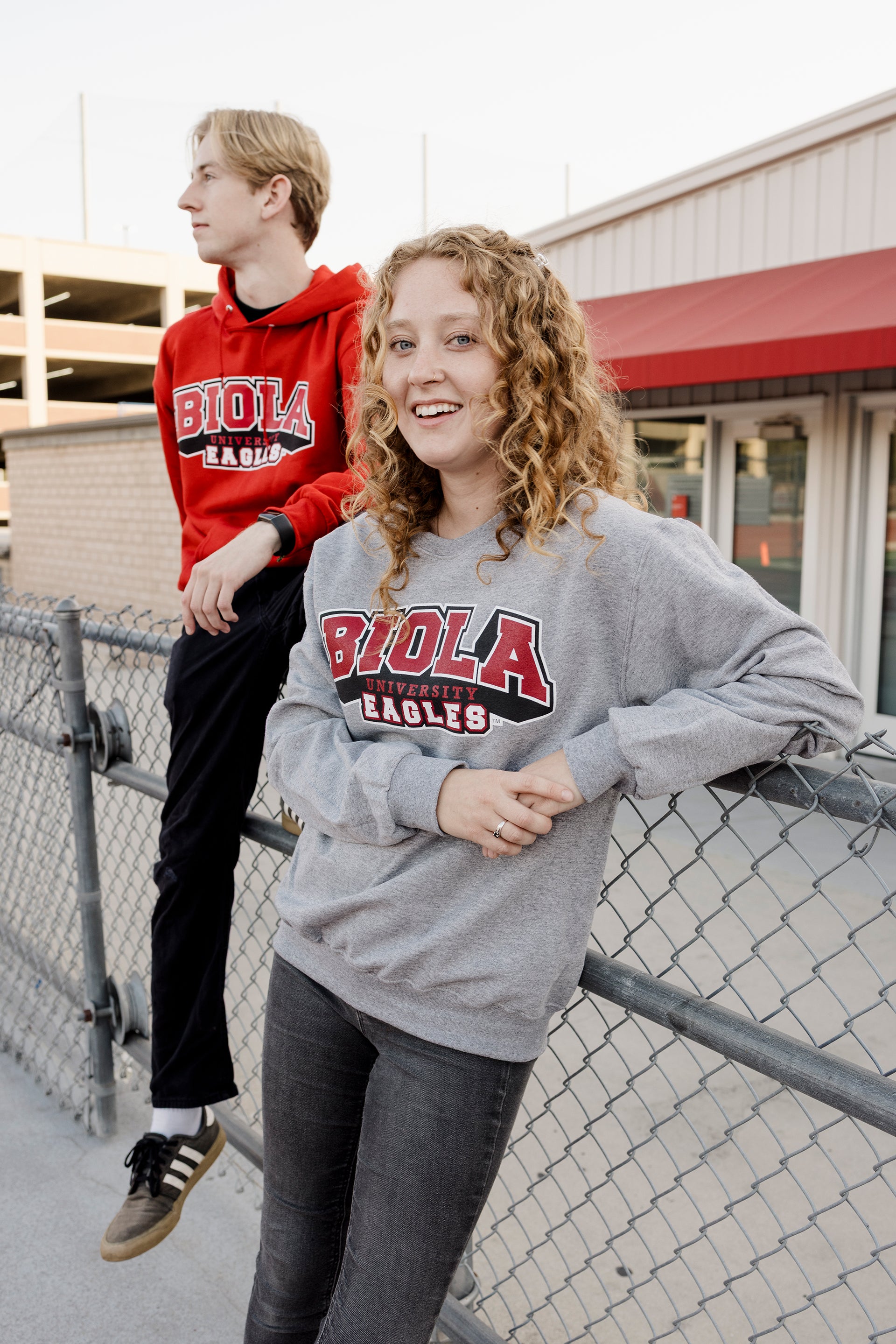 Two people wearing Biola University sweatshirts standing near a chain-link fence.