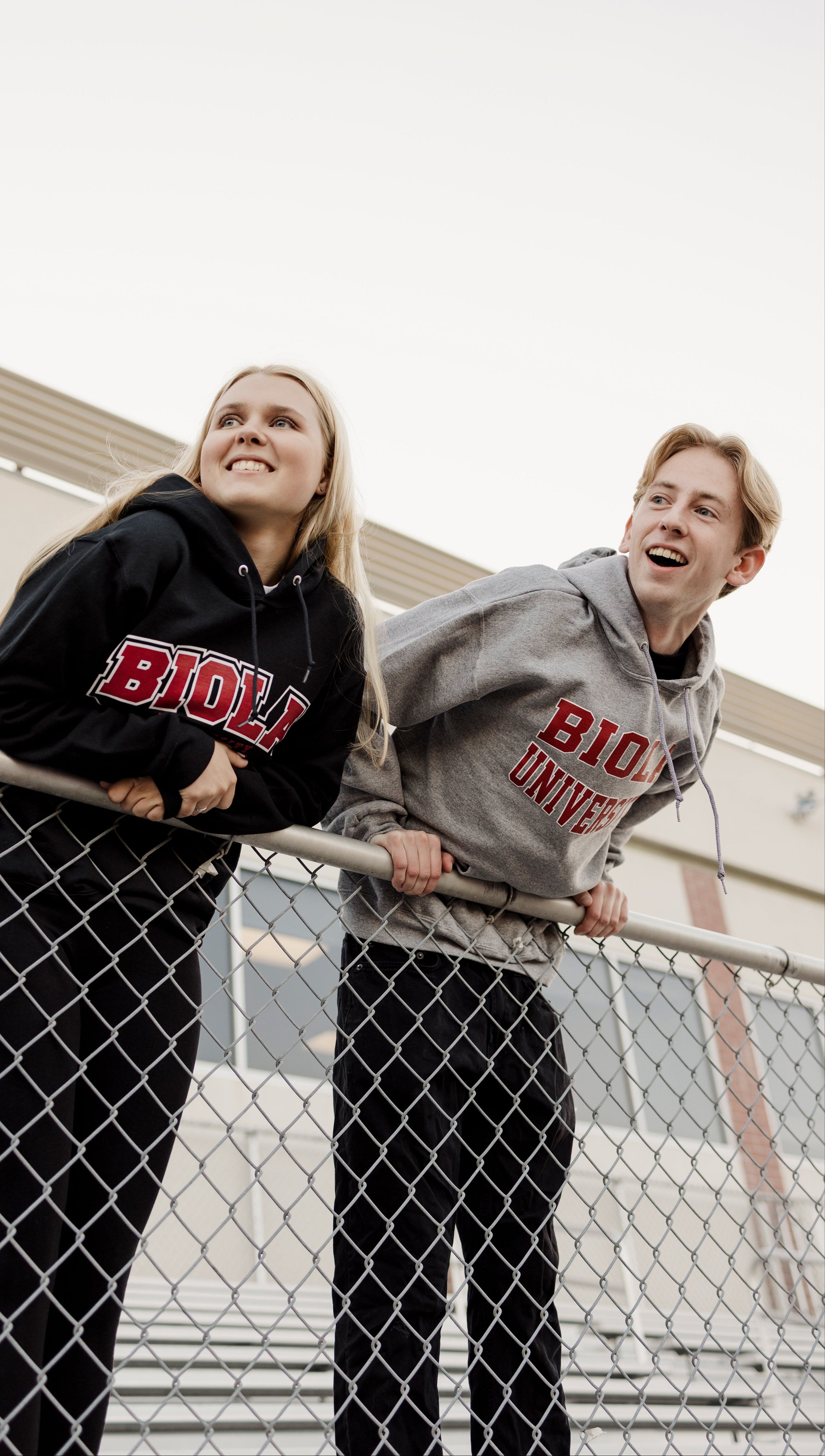 Two people wearing Biola sweatshirts standing behind a chain-link fence.