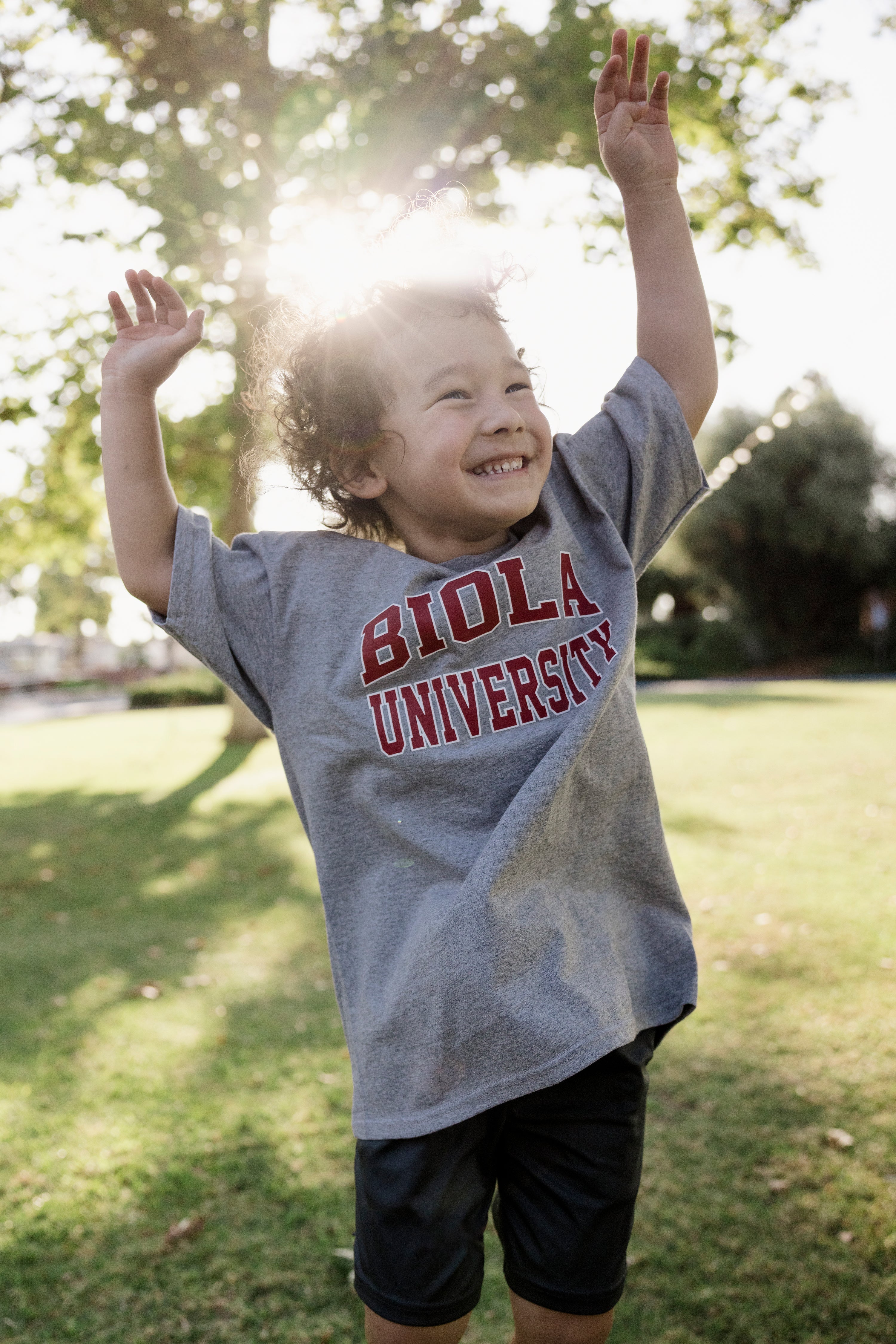 Child wearing a gray 'Biola University' t-shirt with arms raised outdoors.
